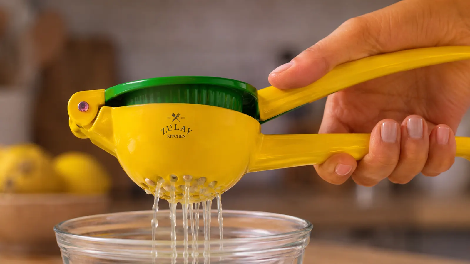 A hand squeezing the handles of a metal lemon squeezer together, with juice draining through the perforated holes into a bowl below while seeds stay trapped inside