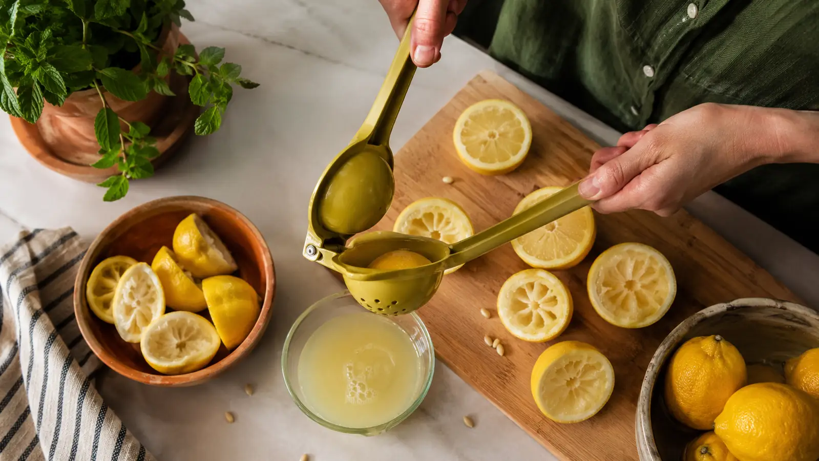 A kitchen counter with three halved lemons, a yellow and green metal lemon squeezer, and a small glass bowl catching fresh juice in morning light