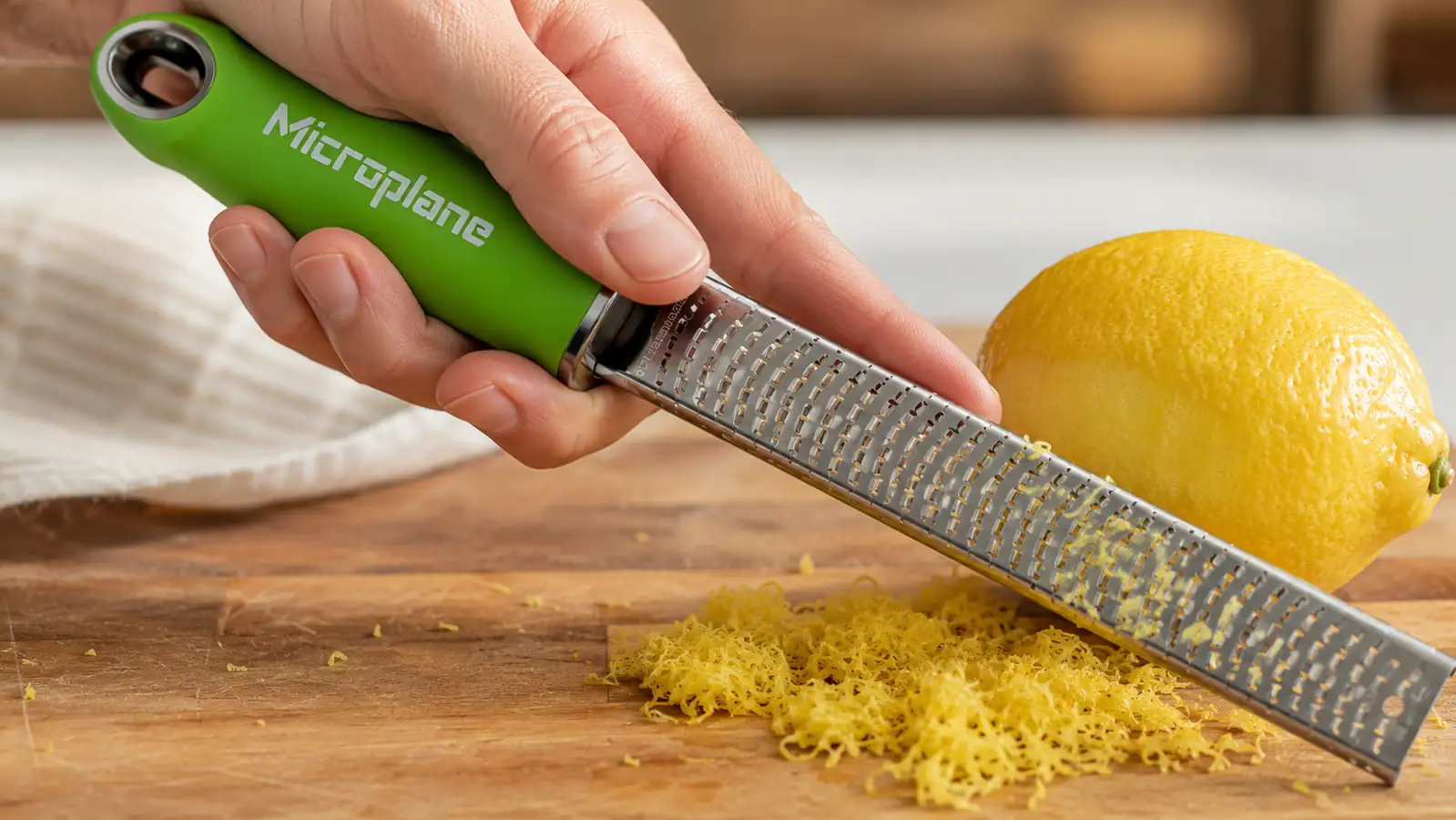 Close-up of a hand dragging the Microplane zester across a lemon at a shallow angle, showing correct zesting technique
