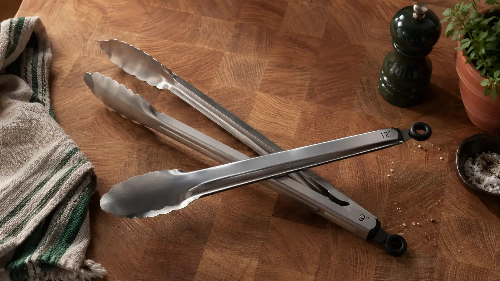 Close-up of stainless steel locking kitchen tongs with silicone tips resting on a wooden cutting board next to fresh herbs