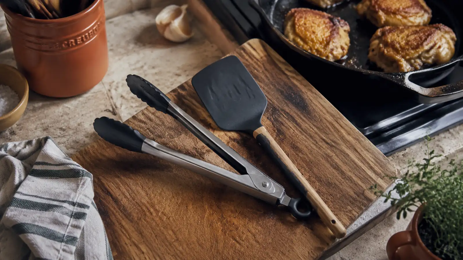 A pair of stainless steel locking kitchen tongs and a silicone spatula crossed on a wood cutting board