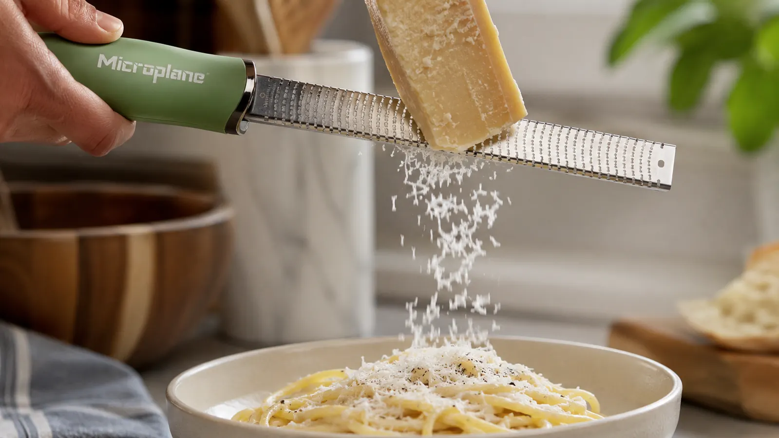 A hand gripping the Microplane zester and dragging it across a wedge of parmesan cheese over a bowl of pasta