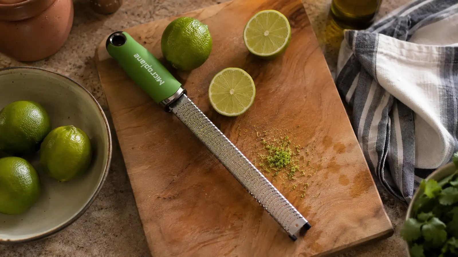 A green-handled Microplane zester resting on a wooden cutting board next to a halved lime and a small pile of fresh lime zest