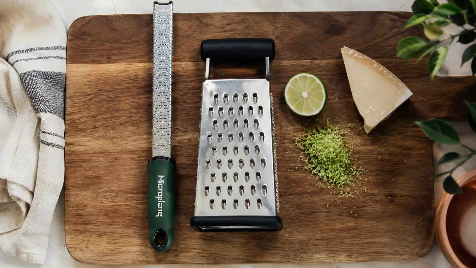 A Microplane zester and a box grater laid side by side on a wooden cutting board with a lime, a wedge of Parmesan, and a small pile of fine citrus zest between them